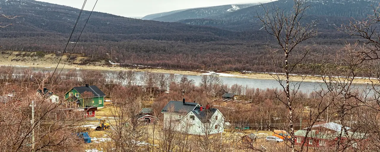 Taloja joen rannalla Utsjoella, taustalla metsäisiä tuntureita.